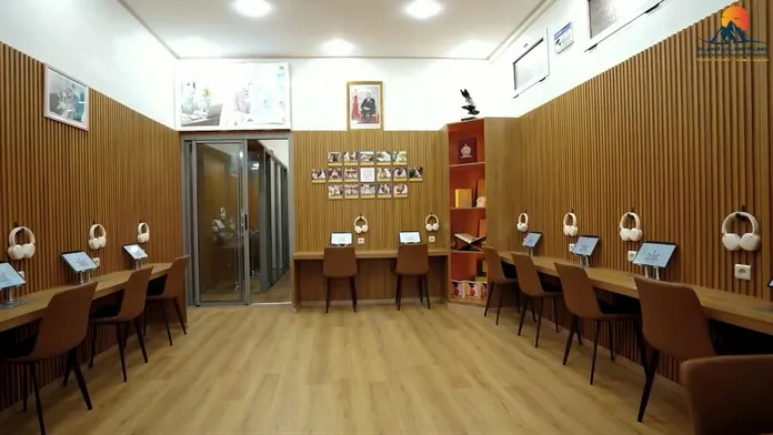 The AI Quranic education room: wood-panelled walls, rows of desks with tablets and headphones, framed works on the far wall.