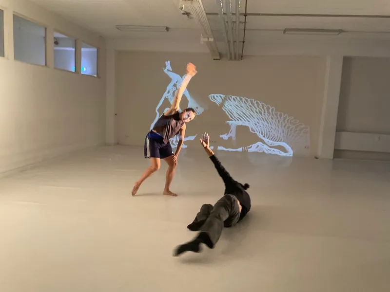 Two dancers in a bright studio in front of a projected line drawing of a large animal.