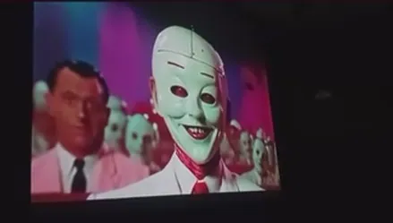 Projected video frame: a grinning white mask among smiling faces.
