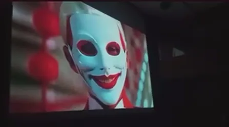 Close-up of a pale mask with a wide red smile on a projection screen.