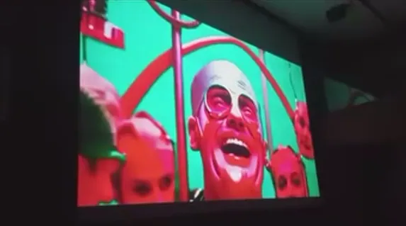 Projected frame of a laughing bald man in round glasses on a green background.