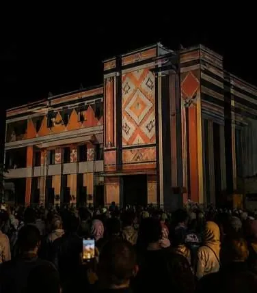 Night façade of a cultural center covered in orange and red geometric projections, crowd in front.