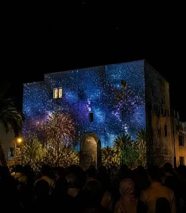 Old stone walls lit with a blue starry-sky projection at night, crowd watching.