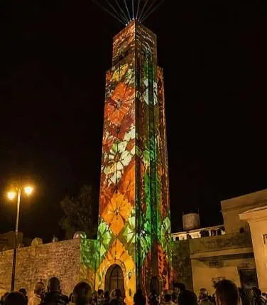 Tall tower covered in orange, red and green patterns, light beams rising from the top.